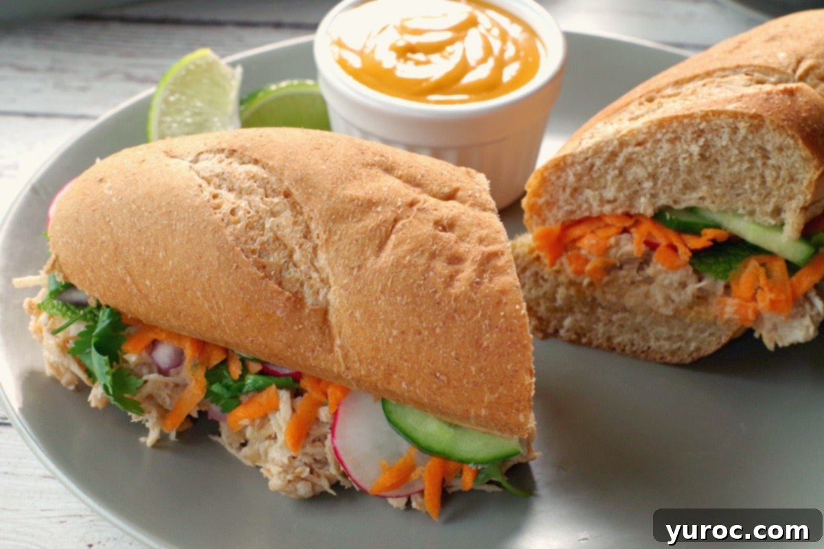 Chicken Vietnamese sub, sliced in half, showing interior layers of chicken and fresh vegetables, with a small white dish of Sriracha mayo and lime wedges on a greenish-grey plate