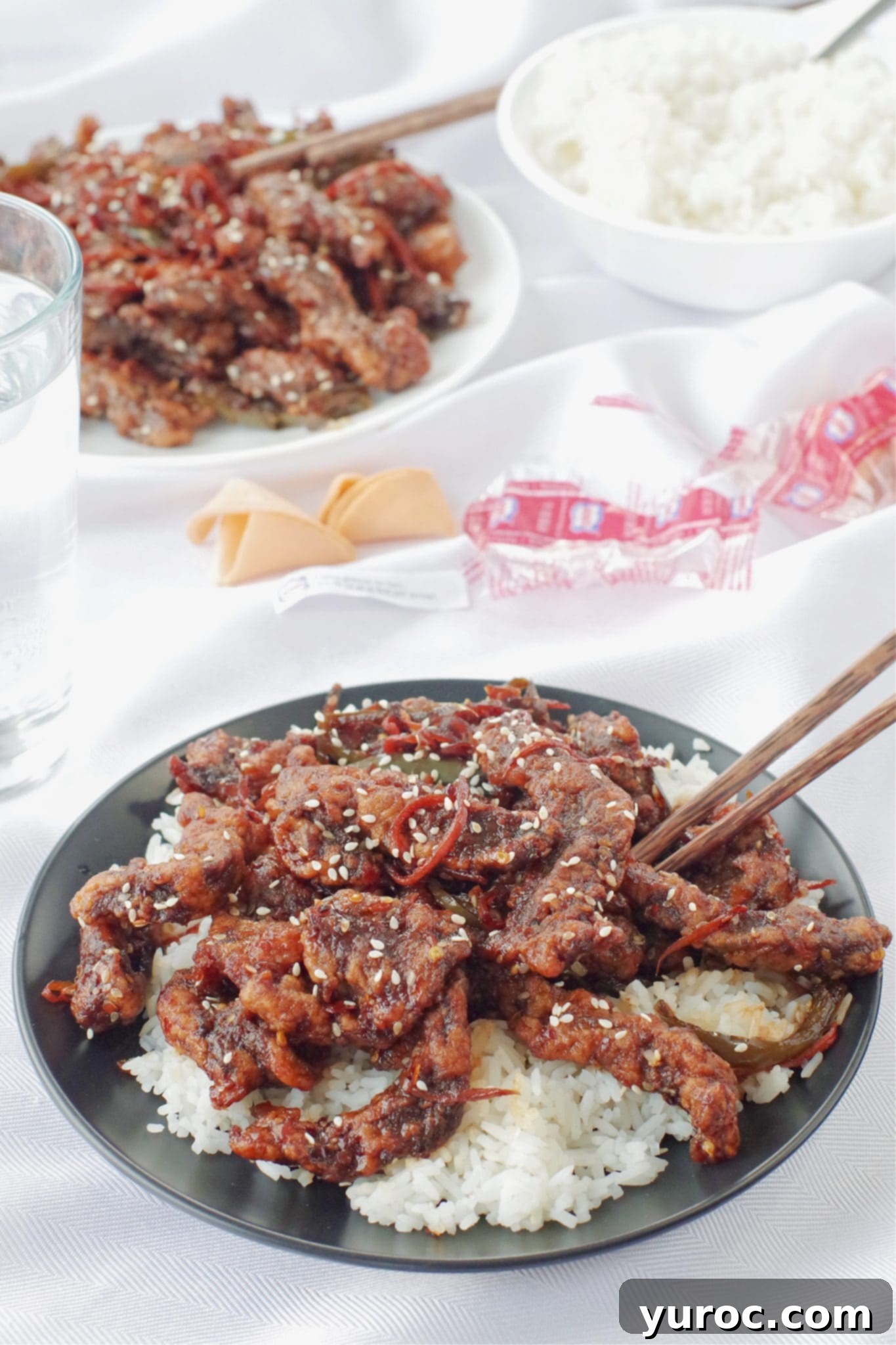 The Ultimate Calgary Ginger Beef 3 ginger beef over rice on black plate, with chopsticks on a white tablecloth, with a dish of ginger beef and rice in the background, as well as fortune cookies