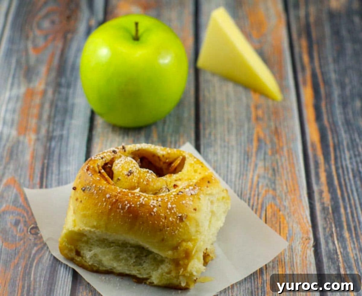 Warm bread machine cinnamon rolls, generously glazed, resting on a blue surface, with a fresh green apple and slices of gruyere cheese blurred in the background, suggesting the key ingredients.