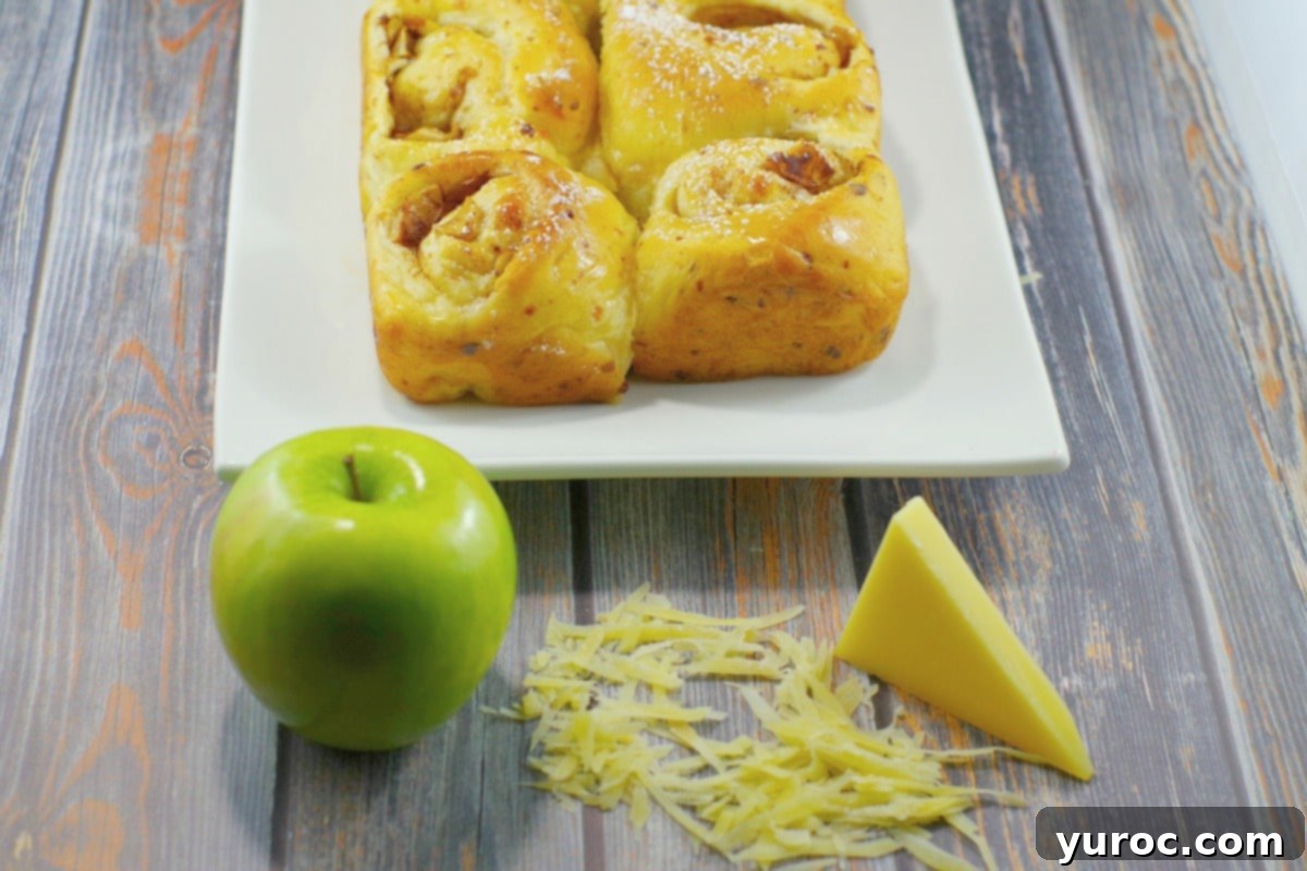 Close-up of baked Apple Pie Gruyere Cinnamon Rolls covered in a creamy glaze, with a sprinkle of powdered sugar, displayed on a wooden surface.