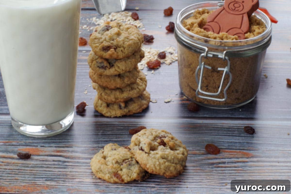 Two soft and chewy oatmeal raisin cookies, one resting partially on the other, with a glass of milk, a stack of more cookies, and a jar of brown sugar in the background.