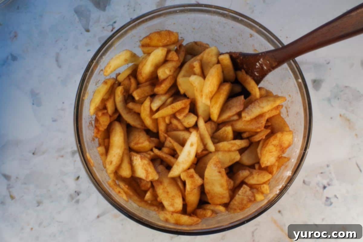Sliced apples mixed with brown sugar cinnamon mixture in large glass bowl with wooden spoon.