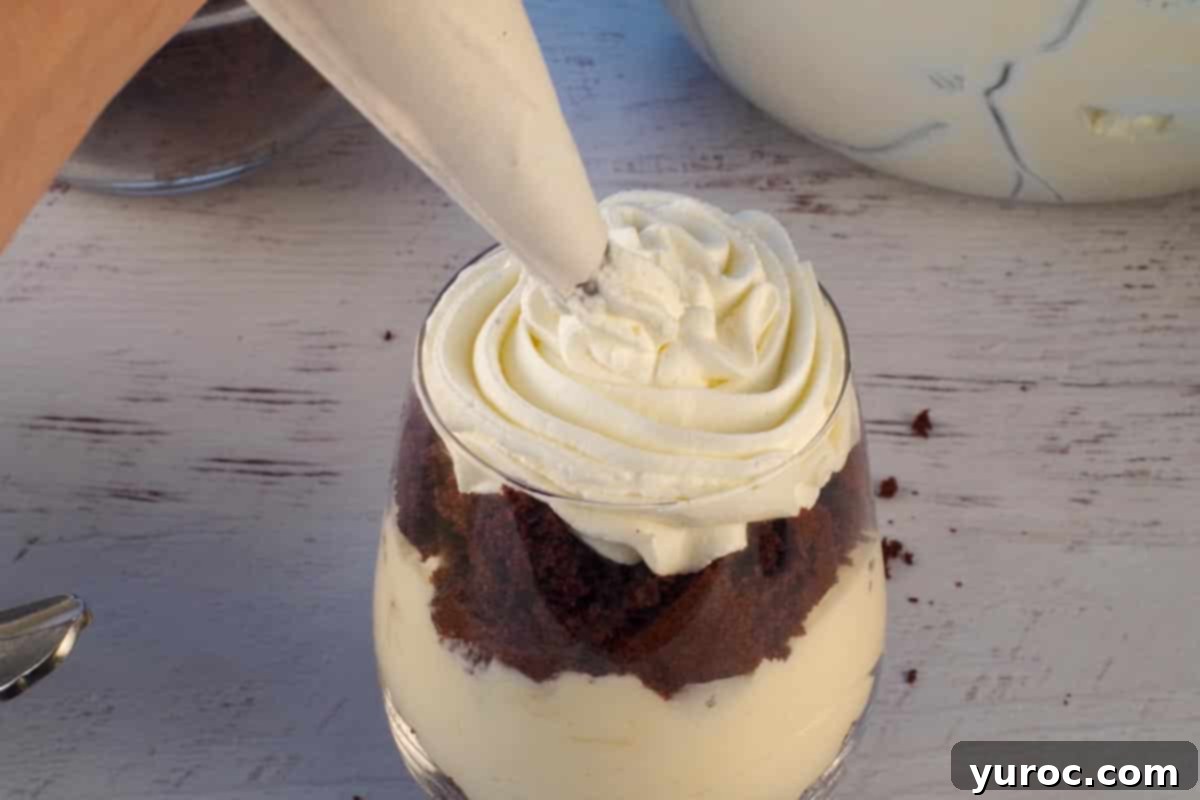 The final step of preparing a mini eggnog trifle, with whipped cream elegantly piped onto the top layer of cake using a pastry bag and a decorative 1M tip, creating a beautiful swirl.