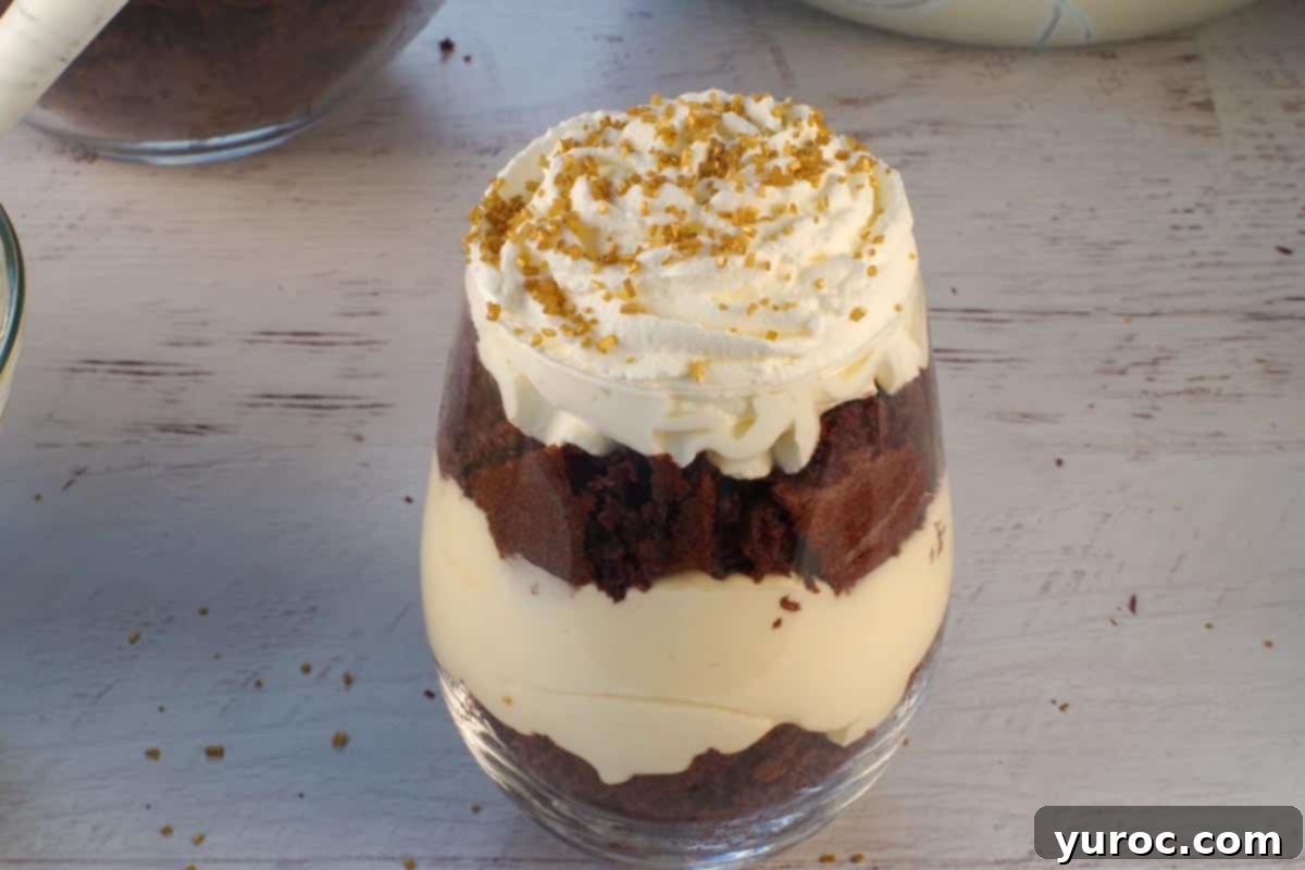 A finished mini eggnog trifle, expertly layered and topped with a swirl of whipped cream, generously sprinkled with sparkling gold edible candy pearls and additional toffee bits, ready to serve.
