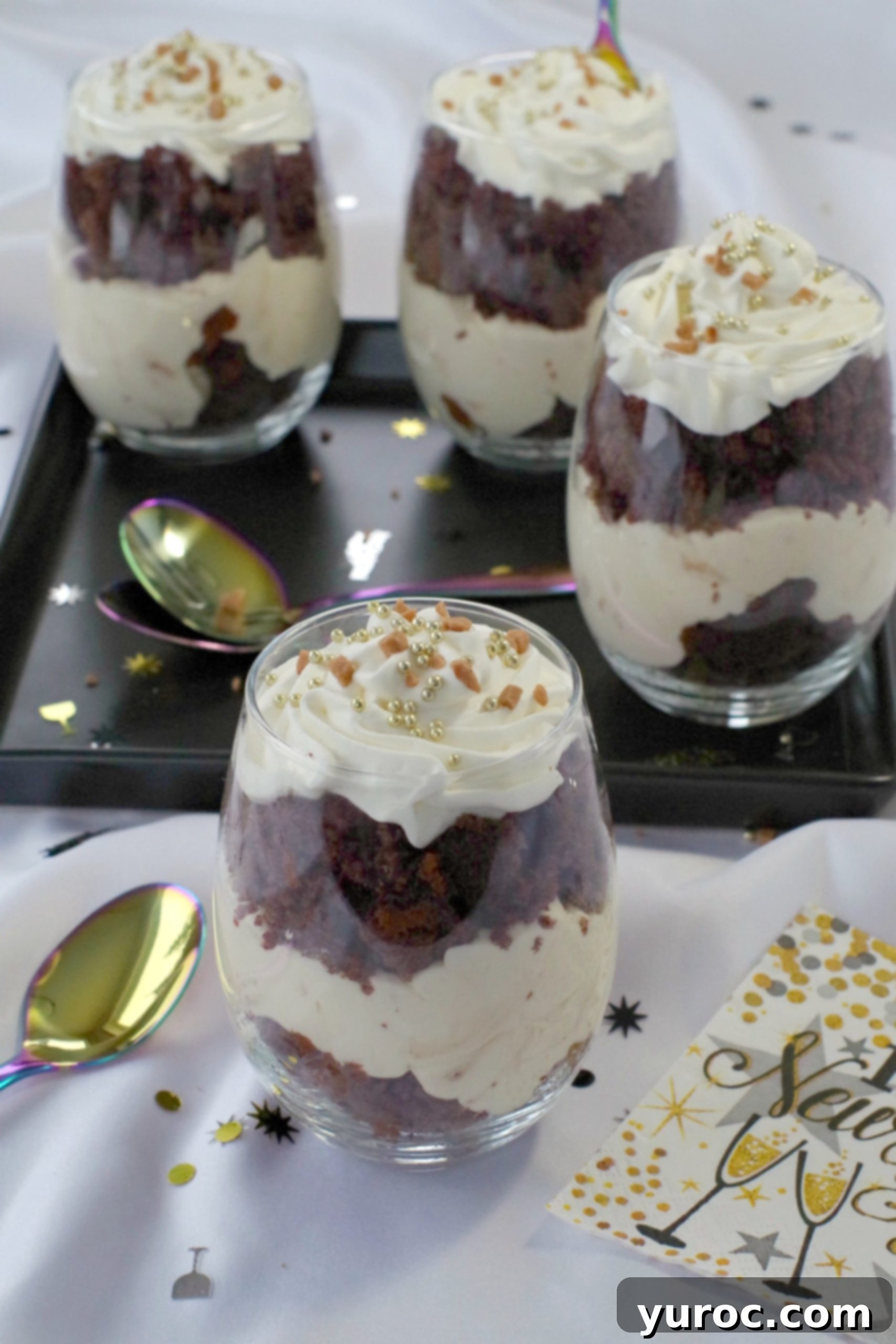 Two vibrant mini eggnog trifles, beautifully garnished with edible gold pearls, positioned on a crisp white background. Elegant New Year's napkins and various holiday decorations surround them, with three more blurred trifles on a black tray in the background, showcasing the dessert's festive appeal.