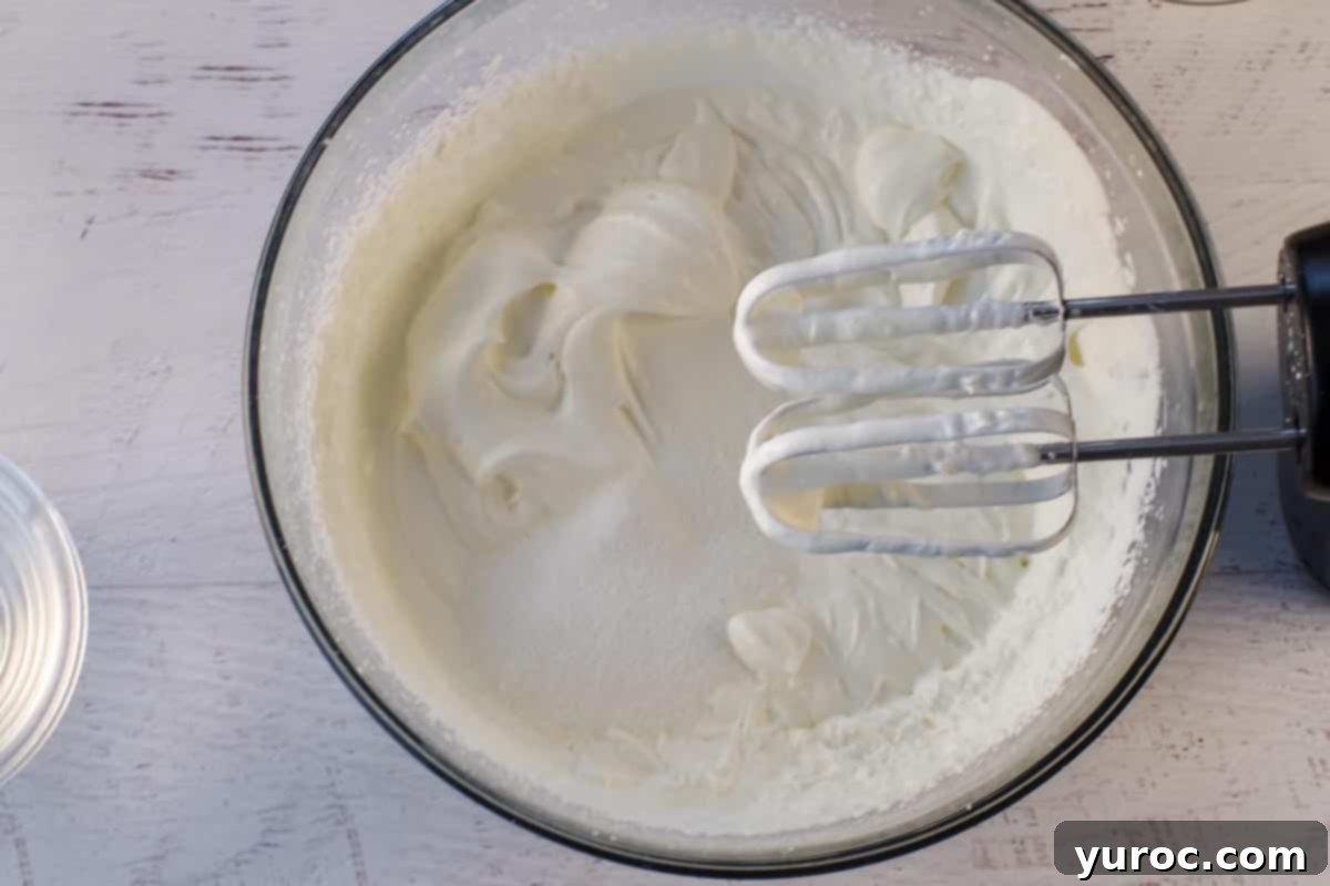Heavy whipping cream being beaten in a large glass bowl, showing the consistency when soft peaks begin to form, with the beaters resting on the rim.