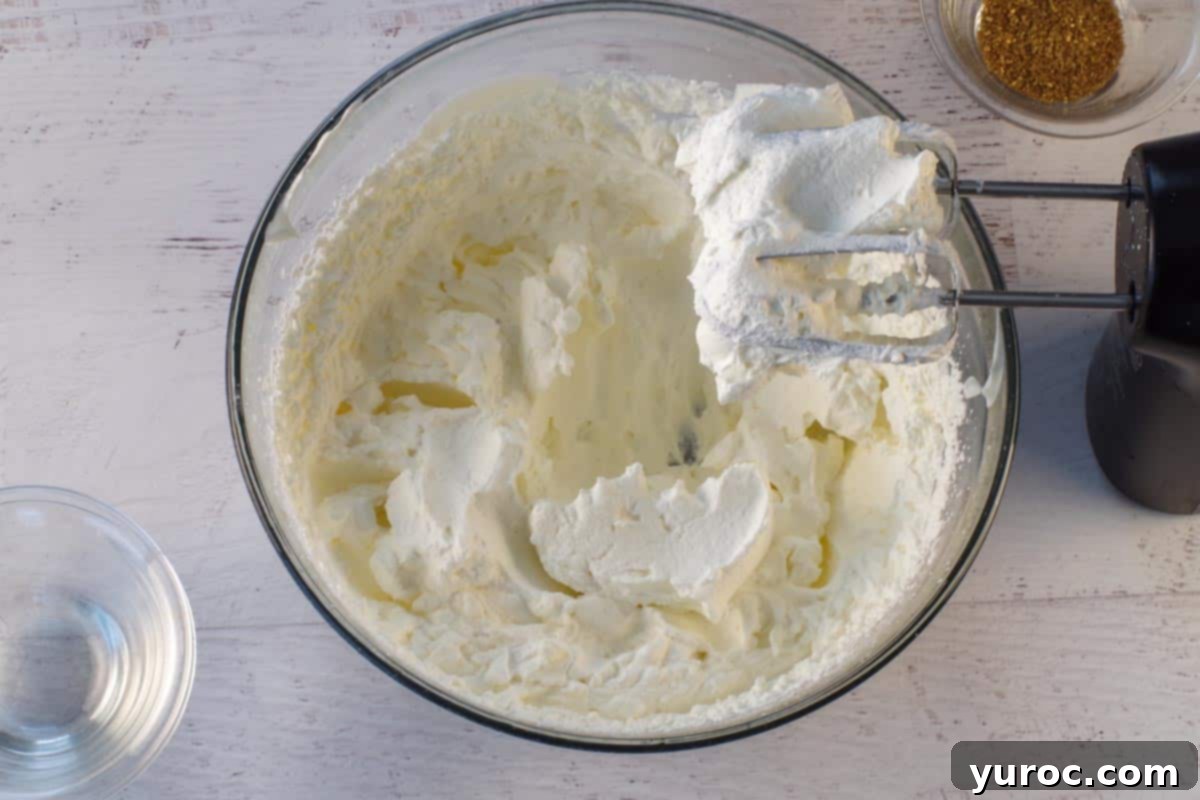 Stiff peaks of whipped cream in a large glass bowl, indicating the perfect consistency for holding shape, with electric beaters positioned above the bowl.