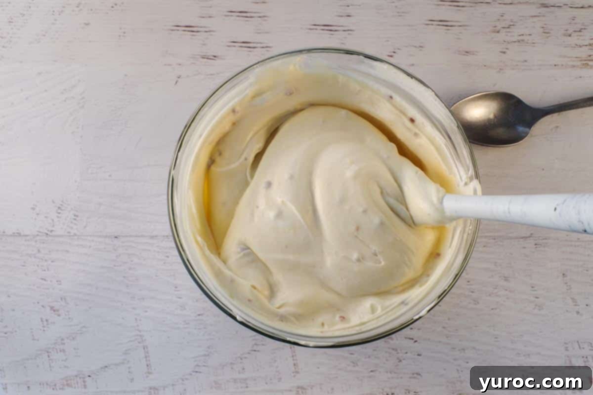 Two cups of freshly whipped cream gently folded into the chilled eggnog pudding mixture in a glass bowl, being stirred with a large spoon to combine, forming the creamy eggnog mousse.