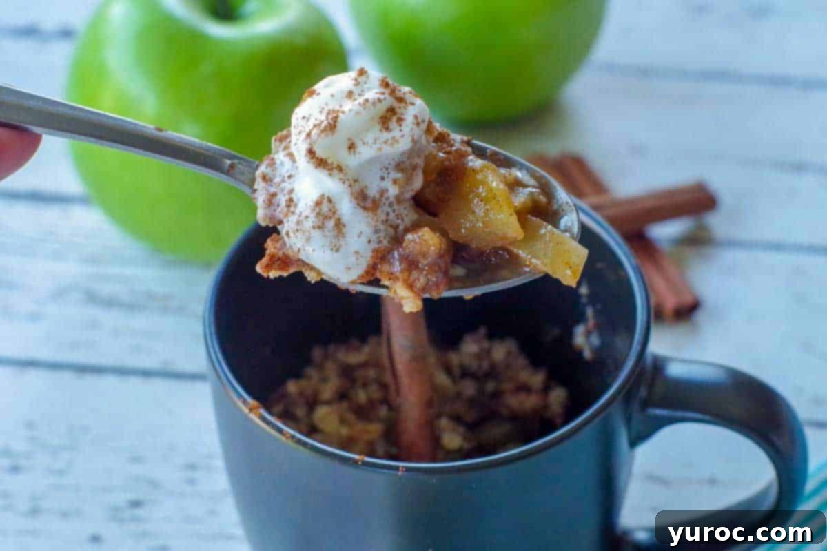 spoon with apple crisp being held up above a black mug apple crisp with 2 green apples and cinnamon sticks in the background