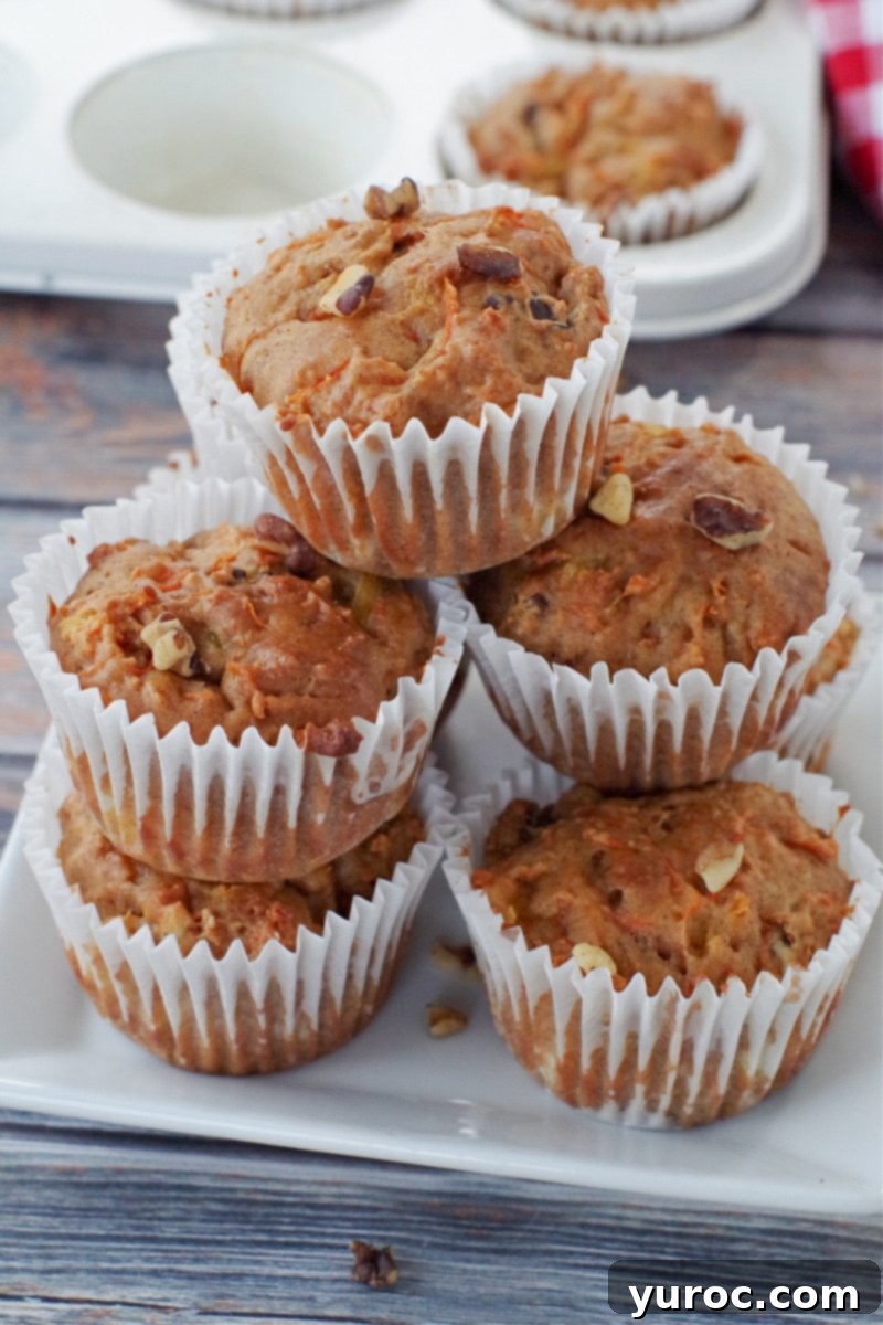 Sunshine Carrot Pineapple Muffins 8 A pyramid of five golden-brown carrot muffins with visible walnuts and pineapple, arranged neatly on a white speckled plate. A muffin tin is visible in the background, out of focus.