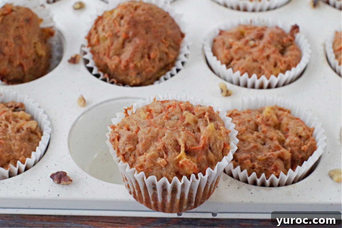 Sunshine Carrot Pineapple Muffins 9 Several healthy carrot muffins nestled in a white speckled muffin tin, with one muffin placed invitingly on top, ready to be enjoyed.