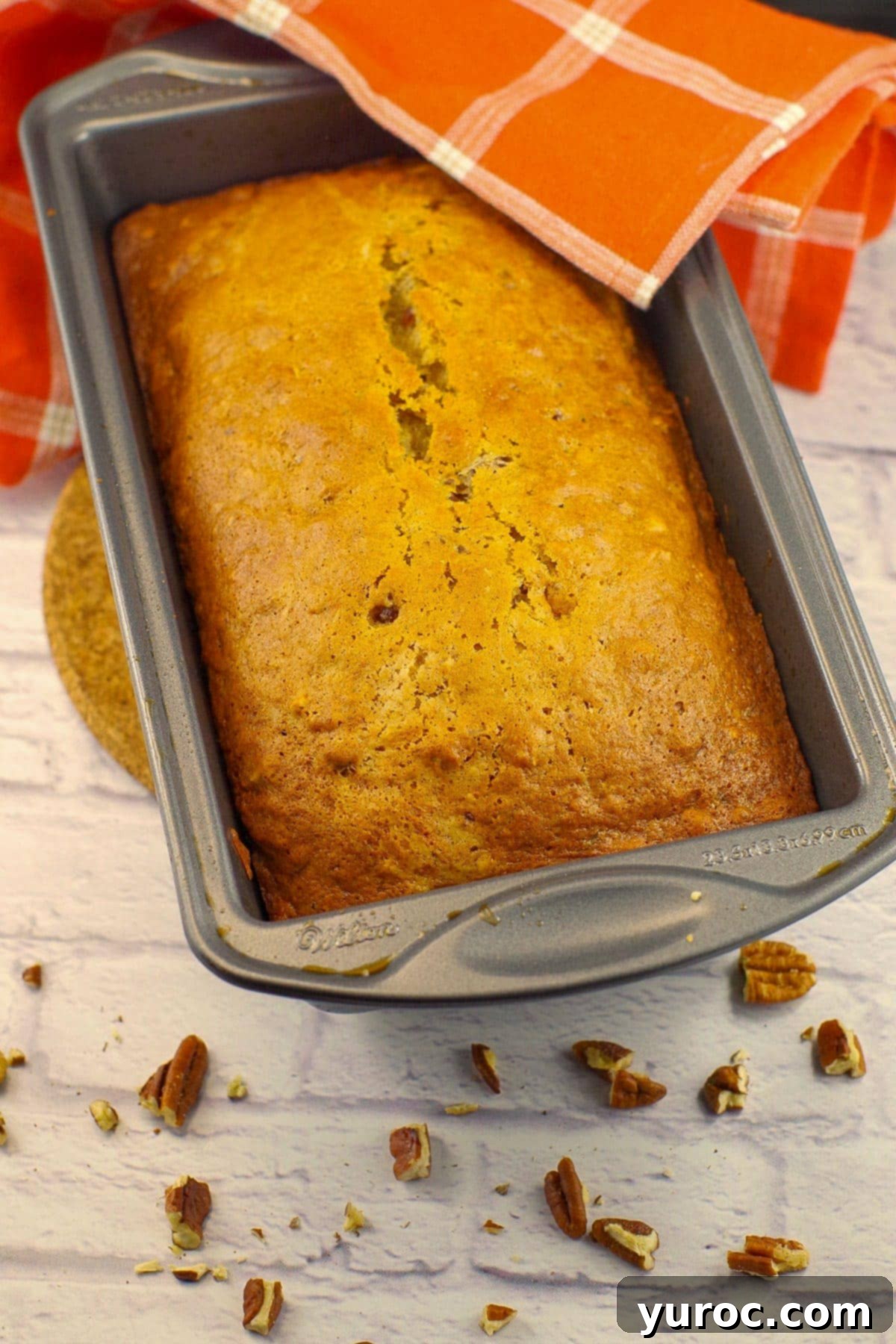 Date loaf in a pan with an orange tea towel and chopped pecans around it and
