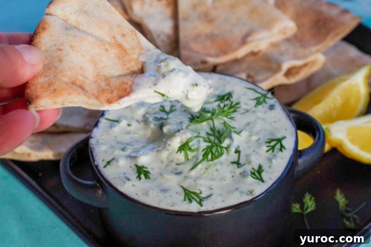 triangle of pita bread being dipped into tzatziki sauce in black bowl on black tray with pita bread in background and lemon wedges
