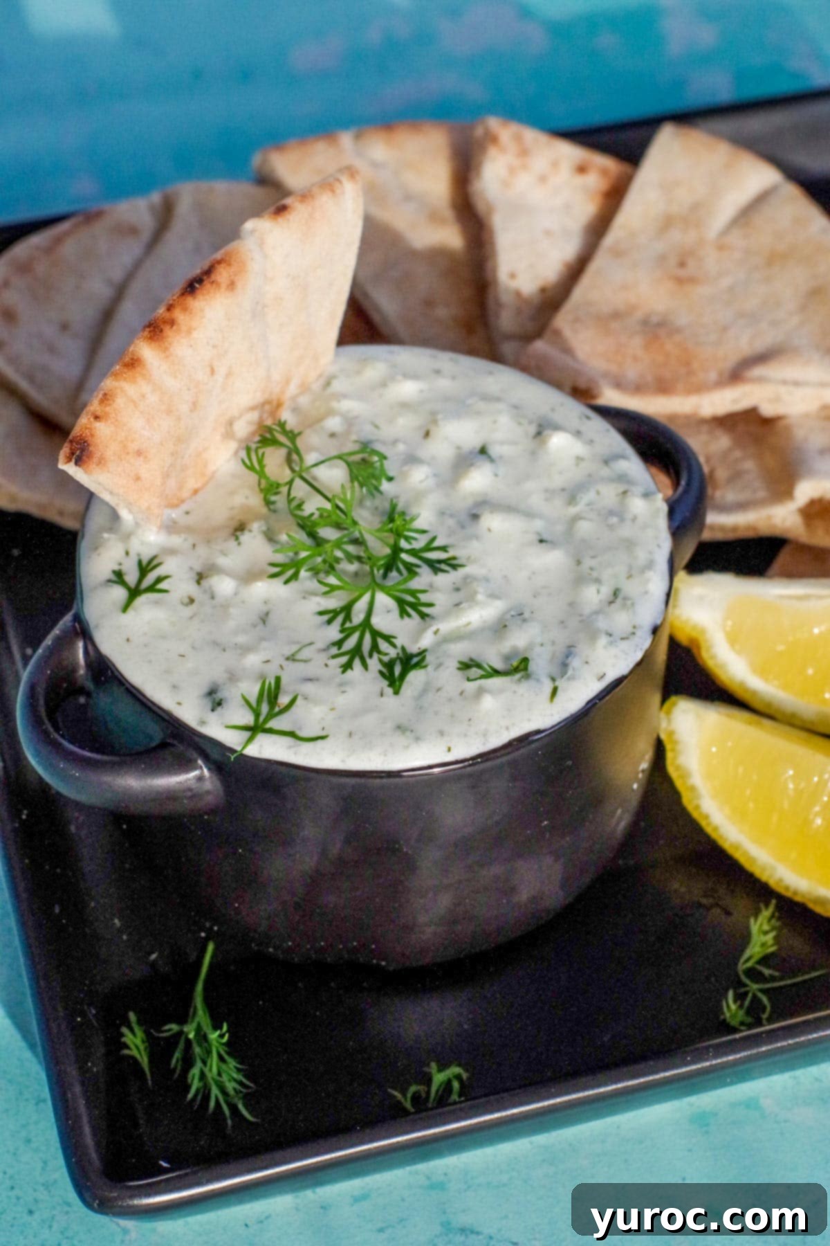 triangle of pita bread being dipped into tzatziki sauce in black bowl on black tray with pita bread in background and lemon wedges