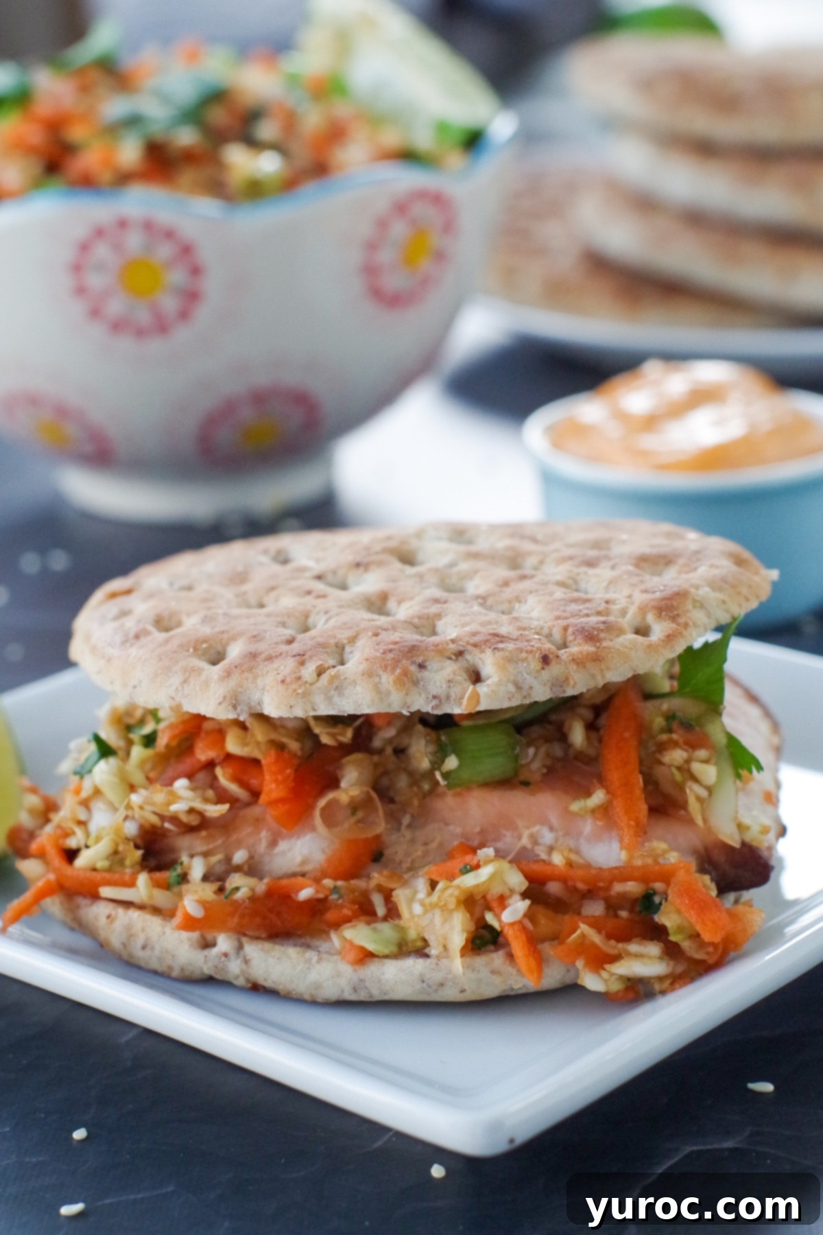 A perfectly assembled air fryer salmon burger resting on a pristine white plate, accompanied by a portion of creamy coleslaw. In the background, fresh burger buns and a bottle of sriracha mayo are ready for more delicious creations.