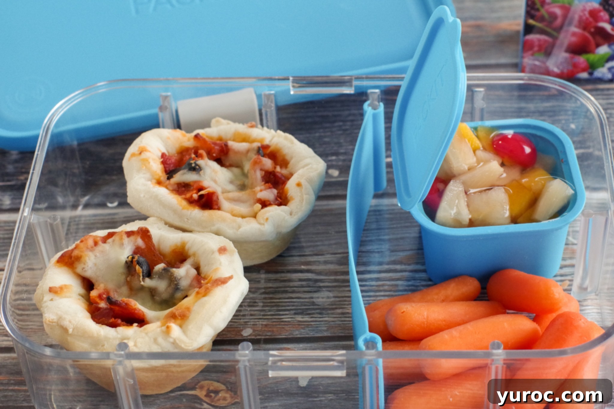 Two pizza cupcakes neatly packed in a clear bento box with blue dividers, accompanied by baby carrots and a small fruit cup