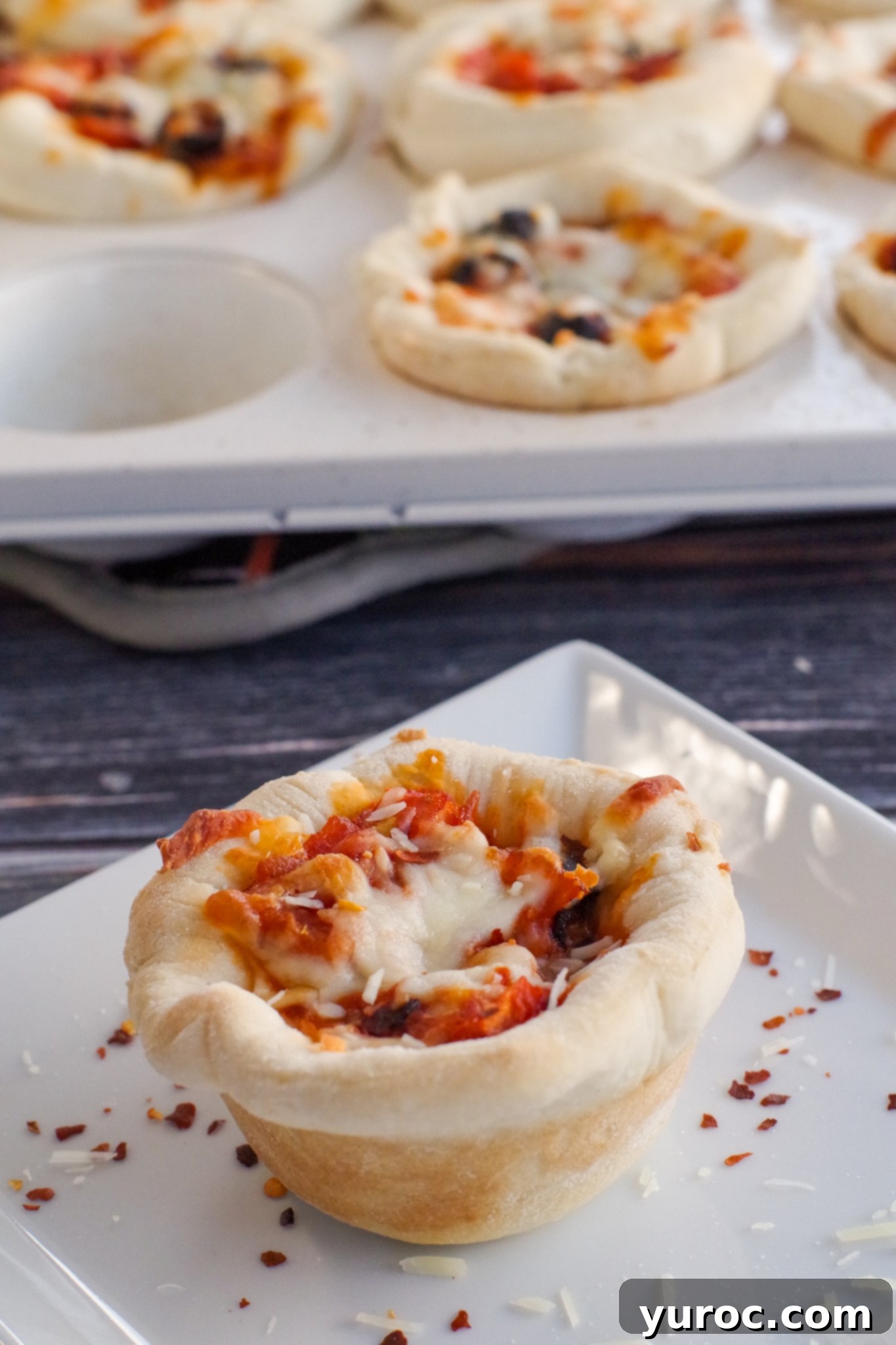 Two savory pizza cupcakes, one on a white plate with crushed red pepper and parmesan cheese, the other in a muffin tin in the background
