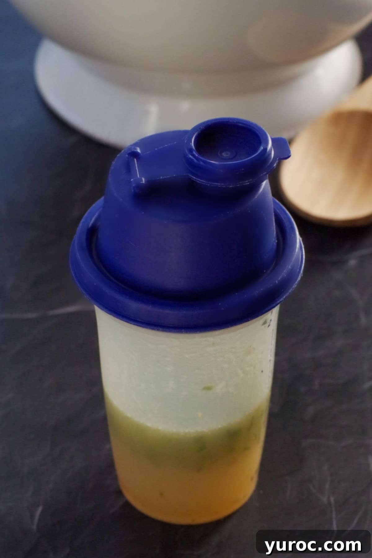 salad dressing in a Tupperware salad shaker with wooden spoon and white bowl in the background