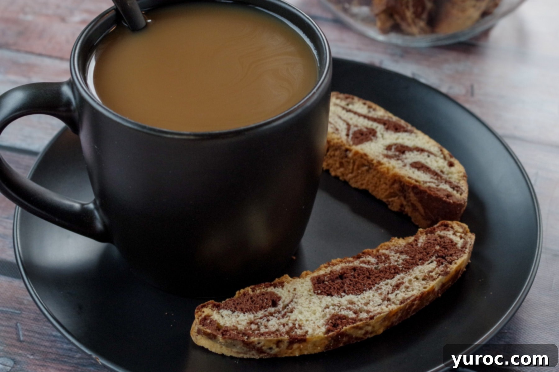 cup of coffee in a black mug on a black plate with 2 pieces of chocolate marble biscotti on the plate and a jar of biscotti in background