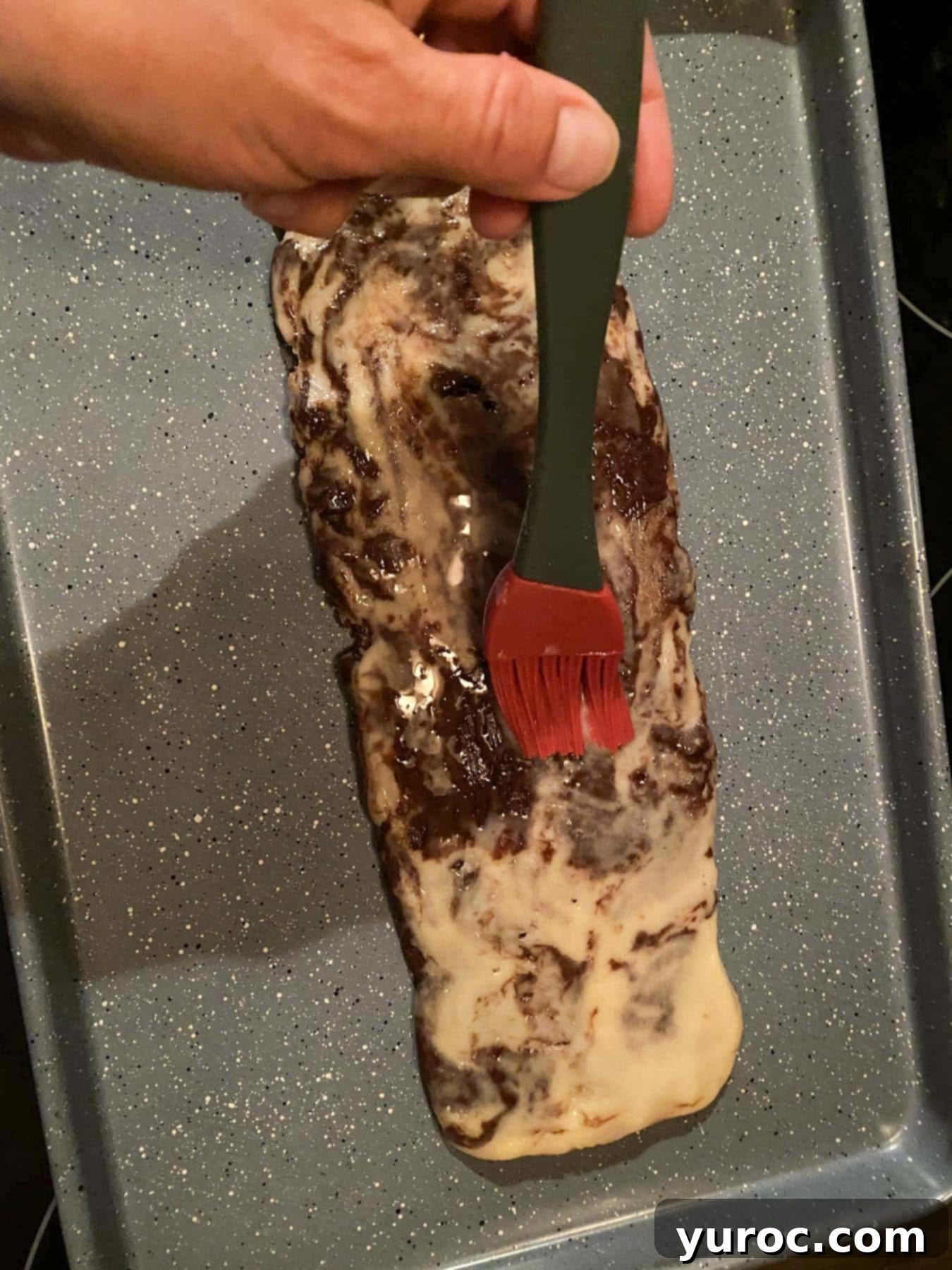 Log of dough on a cookie sheet having an egg wash applied with a pastry brush.