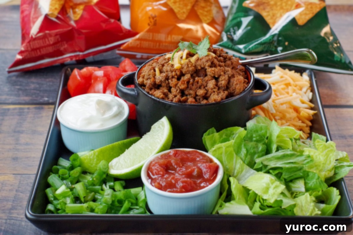 black tray with fixings (lettuce, salsa, sour cream, taco meat, lettuce, cheese, green onions and tomatoes, with bags of tortilla chips in the background