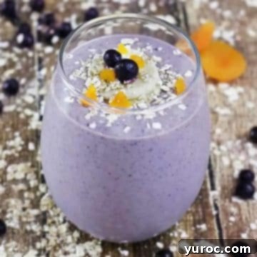 Oatmeal and blueberry smoothie in a glass on a wooden surface, with sprinkles of ingredients around it.
